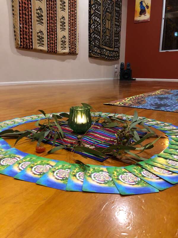 An altar set up in St Kilda. Its a wooden floor with a yoga mat and then an altar of a stripy coloured mat, leaves, a green glass with a lit candle in it surrounded by oracle cards. On the wall behind it you can see African wall hangings.