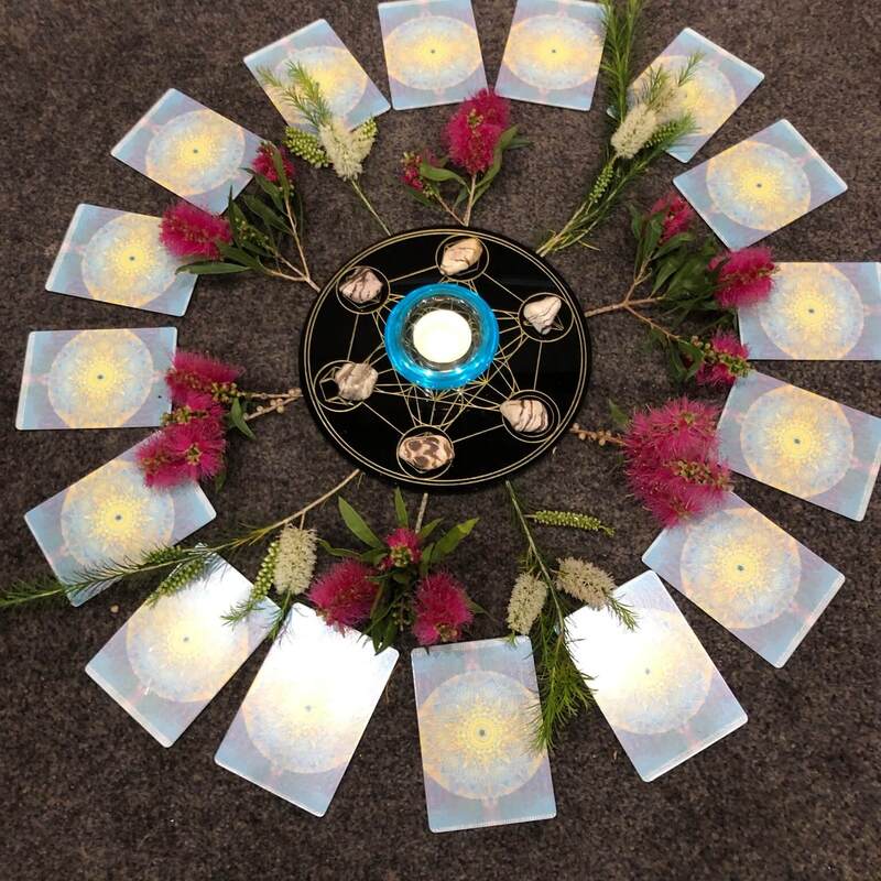 A Qoya class set up. There is a carpeted floor with a black disk in the middle with gold markings on it. There's a blue glass with a tealight candle in it surrounded by crystals, leaves and bottlebrush flowers and oracle cards. 