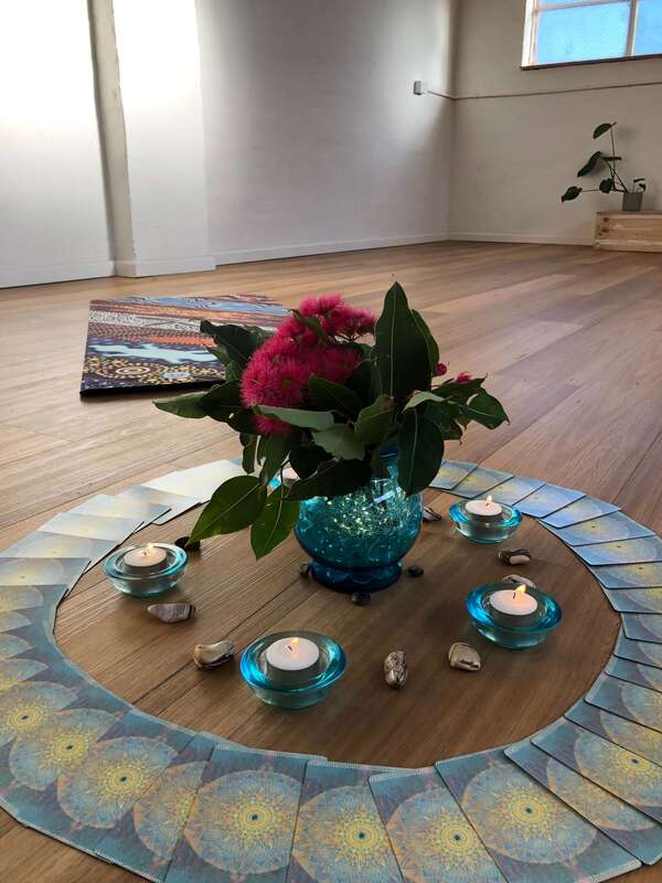 A Qoya class set up. A wooden floor with a yoga mat in the background. There's a blue vase with gum leaves and flowers in it surrounded by tealight candles, crystals and oracle cards. 