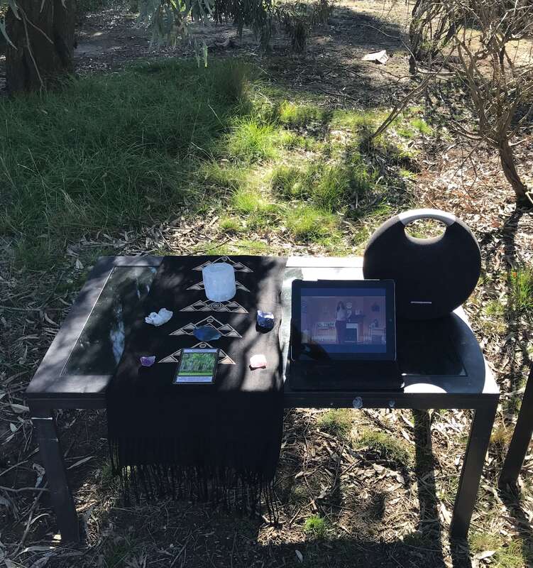 A small table outside set up for a zoom Qoya class for adults. There's a speaker, Ipad as well as a scarf with crystals and oracle cards. 